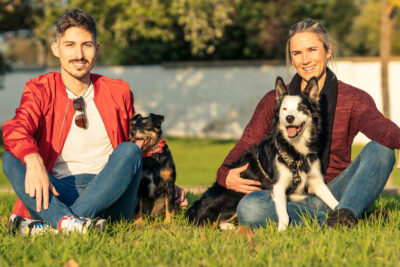 Portrait of two friends petting dogs while looking at the camera sitting on a park