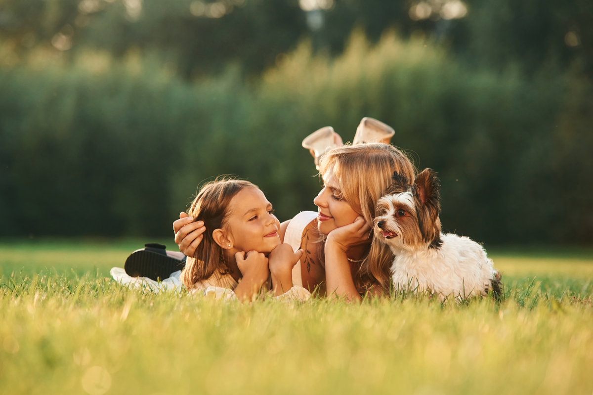 Relaxing, lying down. Mother with her daughter and cute dog are on the field outdoors.